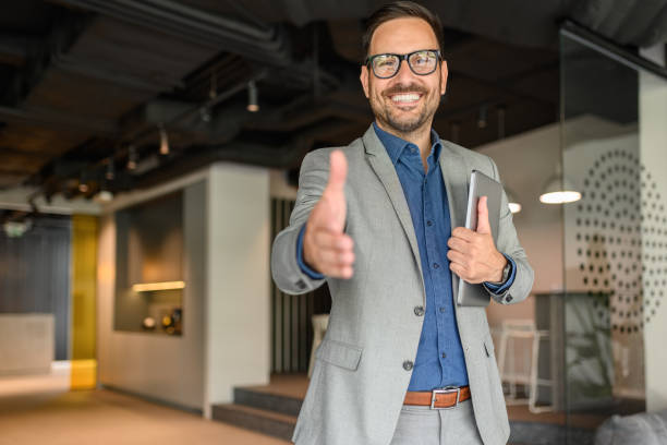 Handsome professional with laptop smiling and offering handshake while welcoming client in office