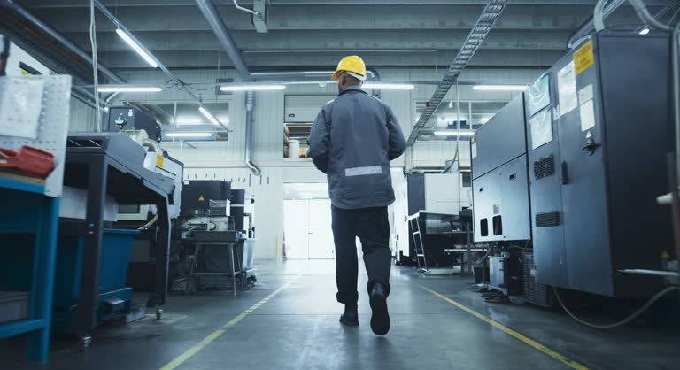 Black Male Worker in a Yellow Hard Hat Walking Through a Modern, Well-lit Factory, Inspecting Advanced Machinery, Checking Equipment, Working in Production Industry. Footage From the Back