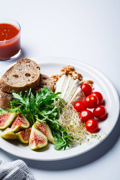 Breakfast plate with bread, tomatoes, brie cheese, arugula, nuts and figs on white background.