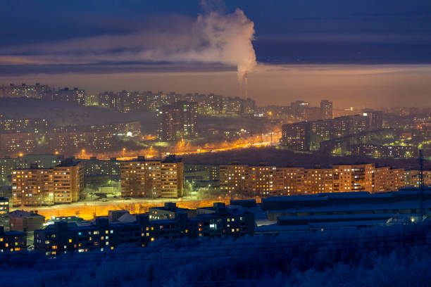 A foggy evening blankets the city, with lights from high-rise windows glowing warmly against the cold winter atmosphere. Murmansk