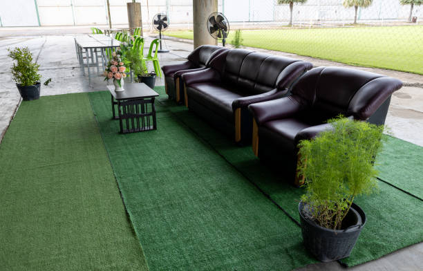 Three brown sofa seats are set up on green artificial grass that is spread out on the concrete floor of the foyer, providing a waiting area for performances near a field in the Thai countryside.