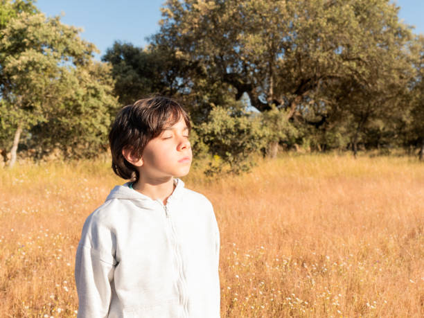 Young boy with eyes closed, basking in sunlight in a serene meadow with trees and dry grass.
