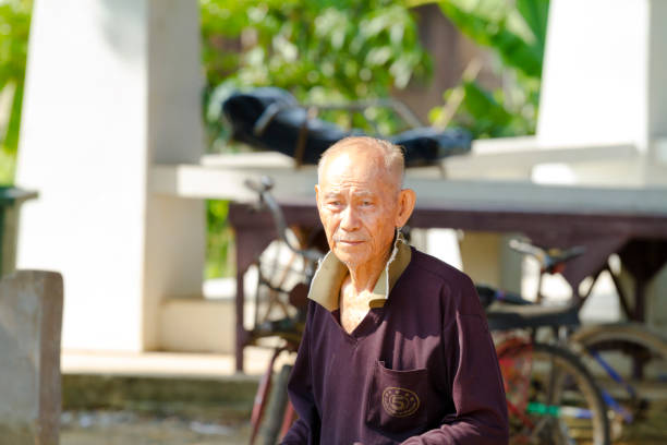 Thai senior man in small village in Kong Krailat District of Sukothai on north western Phitsanulok province captured at morning, Man is standing in street and is looking focused to someone. In background are white columns of a building. There are some old bicycles. Man is wearing a pullover. Man is older person and balding. His eye sight is also suffering and iris has blue rings