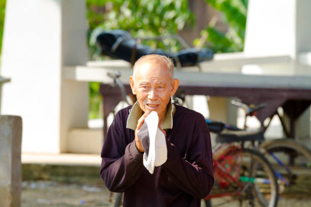 Thai senior man doing wai greeting in small village in Kong Krailat District of Sukothai on north western Phitsanulok province captured at morning, Man is standing in street and is looking focused to someone whom he is greetin with traditional thai wai gesture. In background are white columns of a building. There are some old bicycles. Man is wearing a pullover. Man is older person and balding. His eye sight is also suffering and iris has blue rings