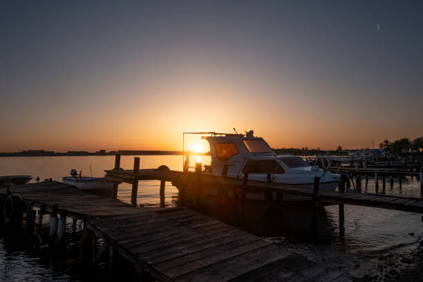 A beautiful silhouette of a boat docked by the pier, captured against the backdrop of a stunning sunset. The sun casts a golden glow, reflecting off the calm waters and highlighting the distant lighthouse. The tranquil scene evokes a sense of peace and calm, perfect for unwinding and appreciating nature's beauty