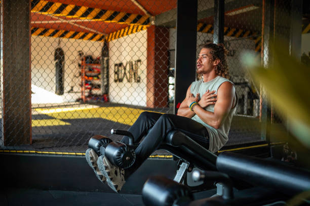 Hispanic male in a gym doing an intense ab workout using a leg raise machine, wearing a sleeveless top and black athletic pants, surrounded by fitness equipment and immersed in focused concentration.