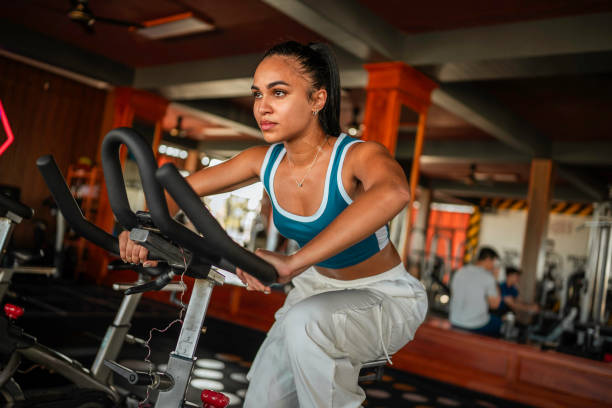 Young Hispanic woman in athletic attire using a stationary bike at a gym. She wears a blue sports bra and white pants, concentrating on her workout amidst gym equipment.
