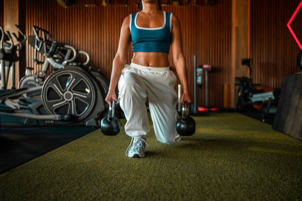 A young adult Hispanic woman, dressed in a teal sports bra and white pants, performs lunges while holding kettlebells in a gym with exercise equipment in the background.