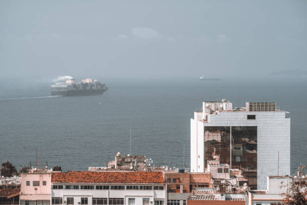 Aerial view of a coastal Rio de Janeiro: a tall building with a red-tiled roof in the foreground and a cargo ship sailing on the horizon in the misty sea background