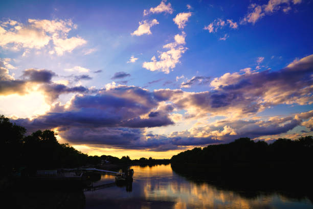 Location: Putney Bridge, London, UK. Putney, West London Sunset, the River Thames, Sunset.The photo is taken from Putney Bridge, looking towards the west-northwest side of London. This perspective captures the expansive view of the River Thames and the skyline of the city, a clean and striking sky is a vibrant blue, dotted with white, fluffy clouds.
