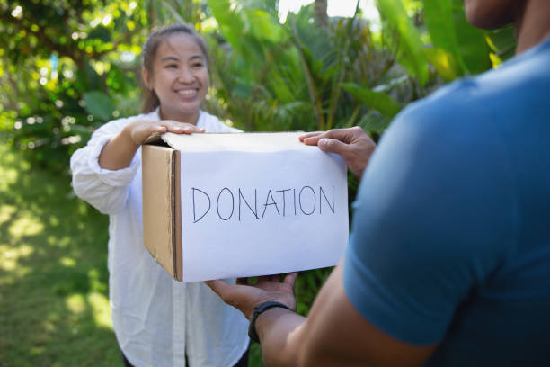 Front shot of a young Asian volunteer giving a donation box to unrecognizable person from charity organization. She's holding a cardboard box with \