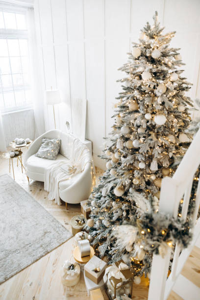 Festive living room featuring white sofa, decorative lights, Christmas tree adorned with ornaments, gold and silver gift boxes, cozy and bright holiday setting, staircase