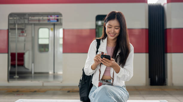 istock Positive Asian female traveler is using her smartphone while sitting on a bench at a train platform. 2163134607