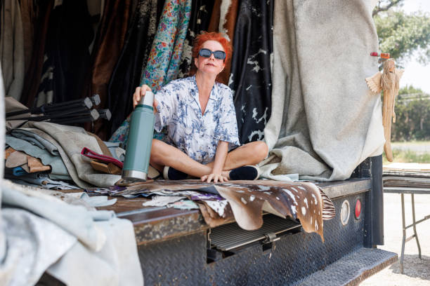 A 53-year-old woman with vibrant red hair exudes confidence as she sits cross-legged in a truck bed filled with cowhides and leather goods. Wearing sunglasses and a patterned blue and white shirt, she manages her small business specializing in unique leather products.