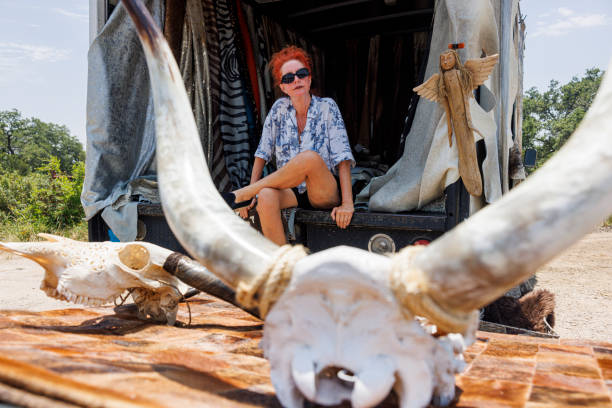 A 53-year-old Texan woman with vibrant red hair sits in the back of a truck filled with cowhides, textiles, and animal skulls with horns. Wearing sunglasses and a blue patterned shirt, she showcases her small business specializing in rustic animal products and western decor.