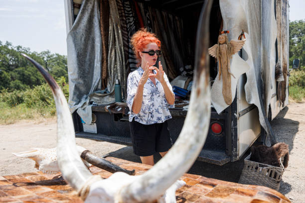 A middle-aged woman with vibrant red hair gestures while arranging cowhides, animal skins, and horns in an open trailer. Parked on the roadside in a grassy rural area, she displays her unique merchandise - exotic leather goods and animal products crafted for a niche market.