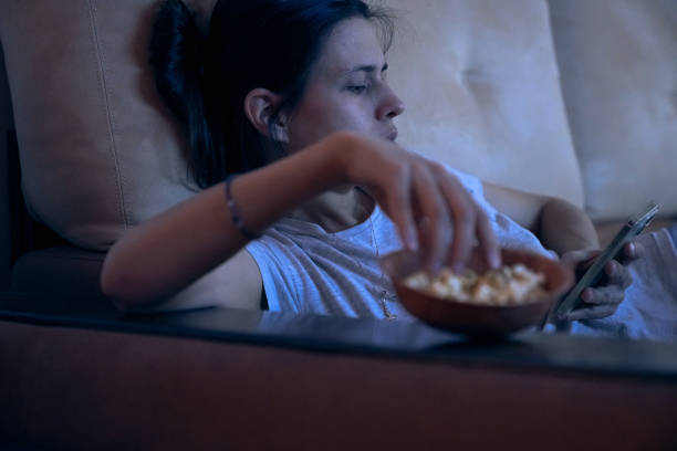 istock Sad woman surfing Internet or chatting using smartphone, lying on sofa and eating popcorn at night. Concept of insomnia, addiction, depression, problems. Background with copy space 2163146859