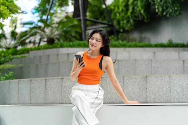 istock Young Asian Woman Looking Away Smiling While Using Phone in Park 2163162036