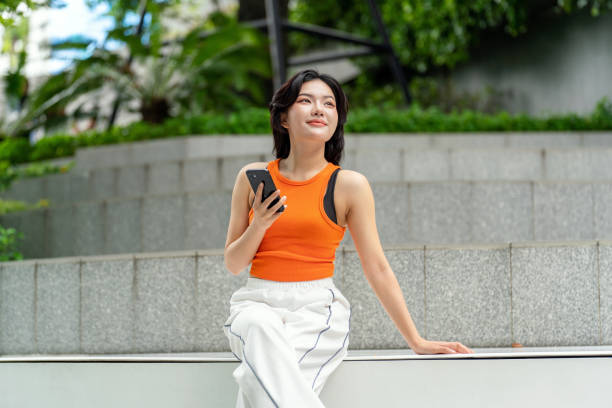 istock Young Asian Woman Looking Away Smiling While Using Phone in Park 2163162080