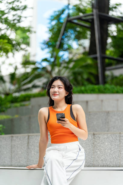 istock Young Asian Woman Looking Away Smiling While Using Phone in Park 2163162157