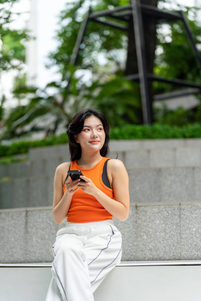 istock Young Asian Woman Looking Away Smiling While Using Phone in Park 2163162228