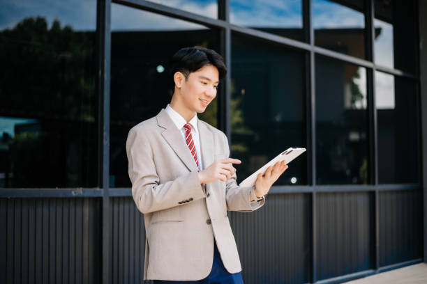 Confident Asian man with a smile standing holding notepad and tablet at out side office. big city on business center