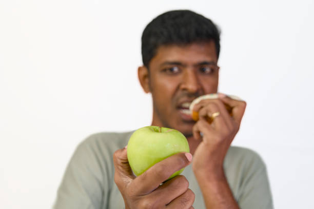 Man deciding between a donut and a fresh green apple, a relatable struggle between healthy and unhealthy food choices.