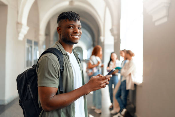 istock Smiling Student Using Smartphone in University Hallway with Friends in Background 2163169778