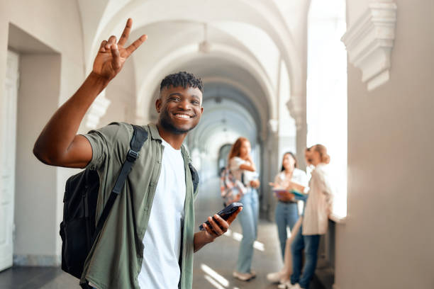 istock A young man with a backpack is smiling and making a peace sign in a university hall 2163169808