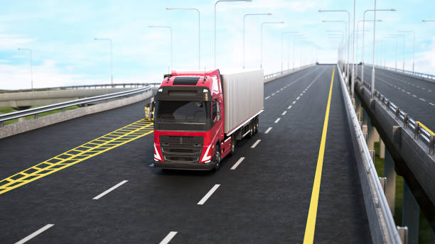 A red semi-truck drives on a highway overpass during the daytime. The truck is in the right lane and is moving forward. The road is asphalt and has a yellow dividing line. There are white lines in the lane. The truck has a white trailer. There are streetlights along the sides of the road.