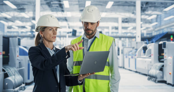 istock Two Professional Heavy Industry Engineers Wearing Hardhats Discussing Factory Work on a Laptop Computer. Caucasian Female Specialist and Male Technician Working On Modern Electronics Production Plant. 2163197976