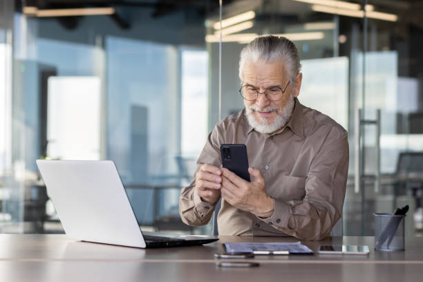istock Senior businessman using smartphone at office desk with laptop 2163201966