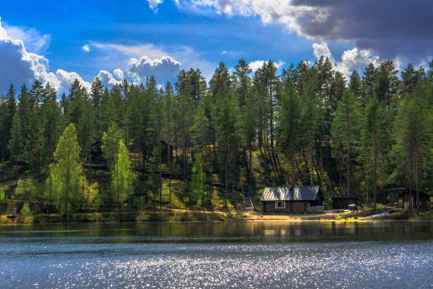 Rokua National Park in northern Finland is known for its unique sand dune landscape, which was shaped by the retreating glaciers of the last Ice Age, In the photo is a view of one of the lakes in the park with a summer house in the opposite shore, with sun creating interesting shadow effects in the nordic pine and birch forest. The first green leaves of springtime have budded in the birch trees. The blue sky with some clouds reflects from the lake