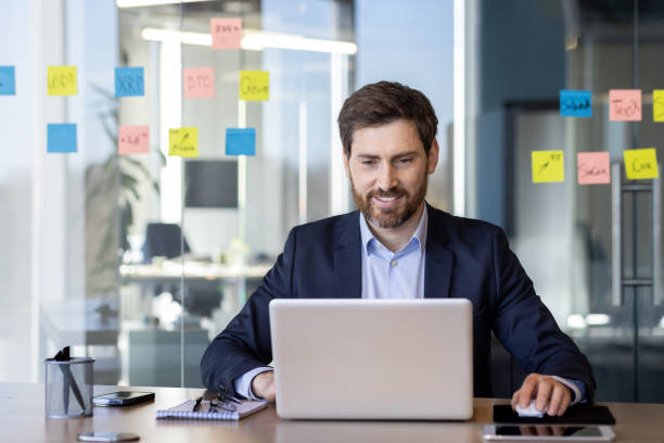 istock Successful smiling man working with laptop inside office, satisfied with results businessman typing on keyboard, mature boss in business suit at workplace 2163215590