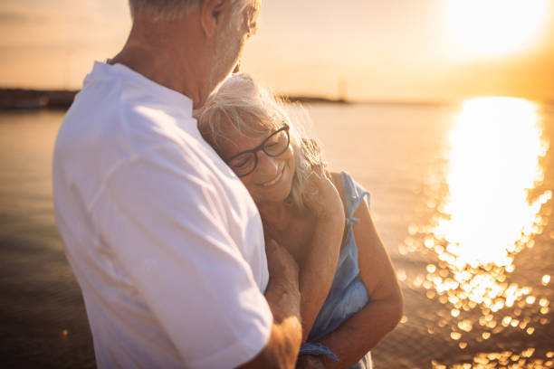 An elderly couple shares a tender embrace on a beach with the sun setting behind them, capturing a heartfelt moment of love, intimacy, and enduring companionship.