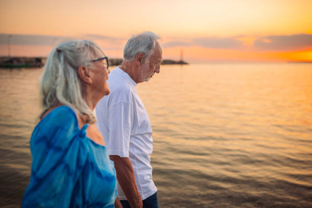 An elderly couple walks along the beach at sunset, enjoying a peaceful moment together with the warm glow of the setting sun illuminating the serene ocean scenery.