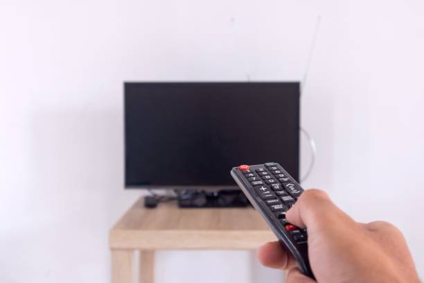 istock Male hand holding TV remote control directed at television on table against white wall 2163220413