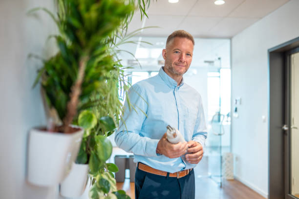 A mid adult Caucasian businessman holds a reusable bottle, smiling in a contemporary office setting, highlighting eco-friendly professional lifestyle.