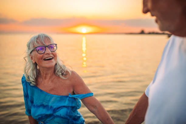 An elderly couple smiling warmly while holding hands and enjoying the tranquility of a sunset at the beach, embodying affection, contentment, and life's simple pleasures.
