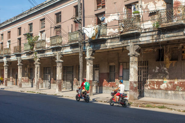 Havana, Cuba - June 25, 2024: Cuban people drive small motorcycle by an old weathered residential building.