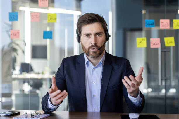 istock Web camera view, man with headset phone inside office talking seriously and focused, mature boss businessman looking at camera, manager on online meeting with subordinates 2163228570