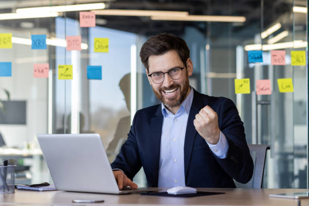 istock Excited businessman celebrating success at laptop in modern office with post-it notes on glass wall 2163235698