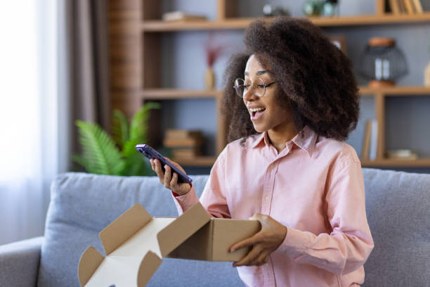 istock Excited woman unboxing package and using smartphone at home 2163265525