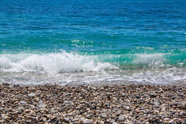 This image captures a tranquil pebble beach with gentle waves lapping against the shore. The clear turquoise waters of the Mediterranean Sea glisten under a bright, sunny sky, showcasing the natural beauty of coastal landscapes.