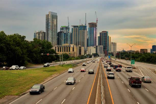 A dynamic view of the Austin, Texas skyline as seen from Interstate 35. The bustling highway is flanked by lush greenery on one side and a cluster of modern skyscrapers under construction on the other, showcasing the city's rapid development. The sky exhibits a soft, overcast glow, adding a serene backdrop to the urban scene. Perfect for social media posts or stock image searches related to urban landscapes, city life, or travel in Texas.