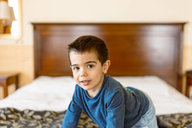 A cheerful young boy wearing a blue shirt is happily playing on a bed in a cozy, well-lit bedroom during the morning time.