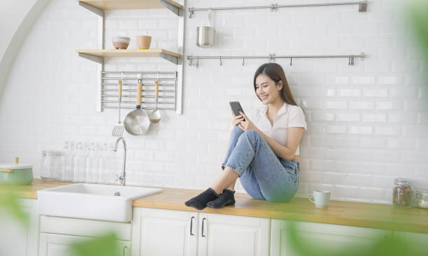 istock Portrait young beautiful asian woman relax sit using mobile cell phone on kitchen counter. Smile happy asian girl drink coffee at table home minimal lifestyle. Freelance small business 2163349682