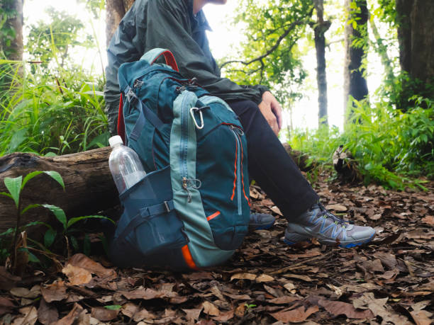 istock Close-up of blue hiking backpack and a man sitting and resting. Backpack has clear plastic water bottle in the side pocket and is surrounded by lush green foliage and fallen leaves. Adventure or camping trip concept. 2163358969