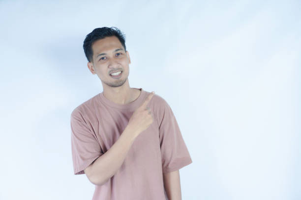 istock An Asian man in a pink shirt points to his side with a surprised and curious expression, as if noticing something unexpected. The plain light blue background emphasizes his animated 2163363537