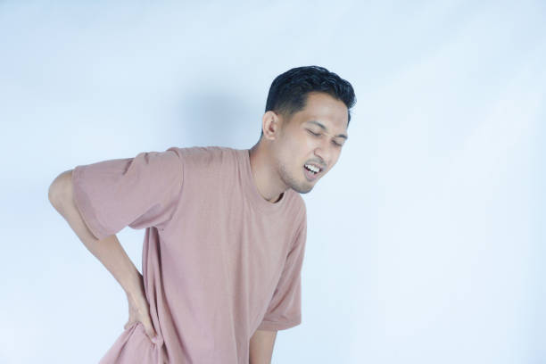 A man in a pink shirt bends over slightly and holds his lower back with a pained expression, indicating back pain or discomfort. The plain light blue background emphasizes his distress